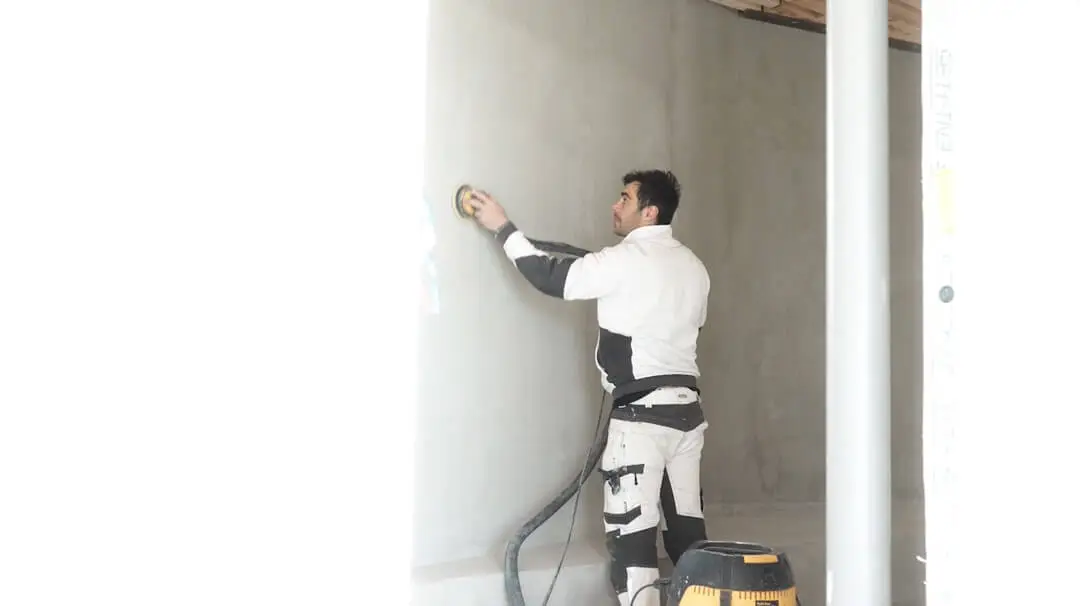 rtisan peintre et décorateur utilisant une ponceuse orbitale reliée à un aspirateur sur un grand mur en béton ciré par Nuances Peinture Décoration.